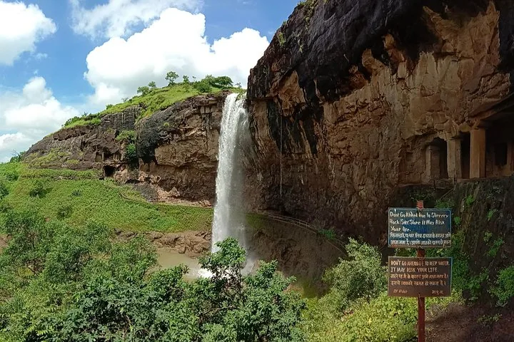 Wasserfall, der neben einem in den Fels gehauenen Höhleneingang mit Warnschildern herabstürzt.